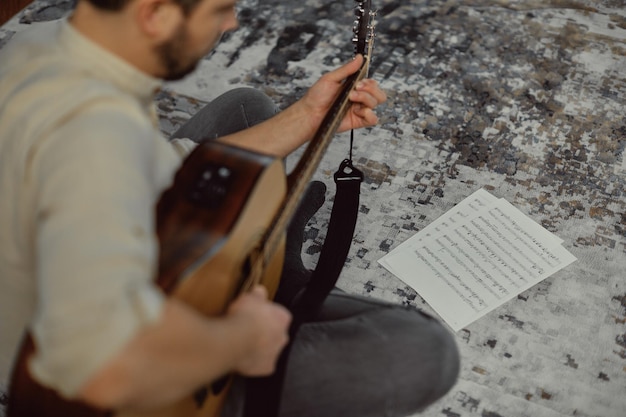 Músico jovem tocando música com violão olhando para partituras sentado no chão