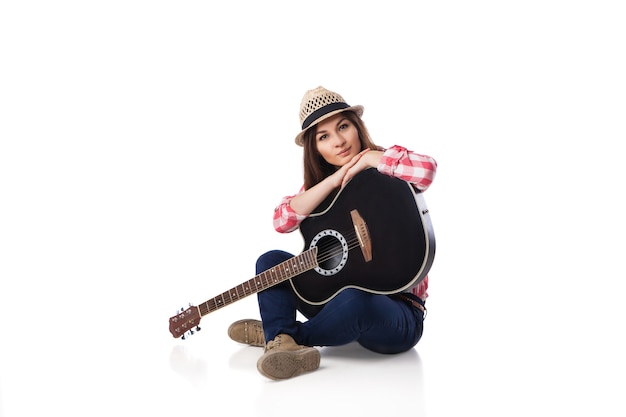 Músico joven con guitarra con camisa y sombrero sentado en el suelo. Fondo blanco.