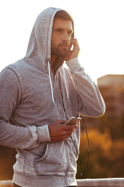 Musik bewegt meine Füße. Hübscher junger Mann mit Kopfhörern, der wegschaut und ein Smartphone hält, während er im Freien steht