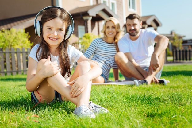 Musik genießen. Entzückendes kleines Mädchen, das auf dem Gras sitzt und die Musik hört, während ihre glücklichen Eltern im Hintergrund sitzen und sie bewundernd ansehen