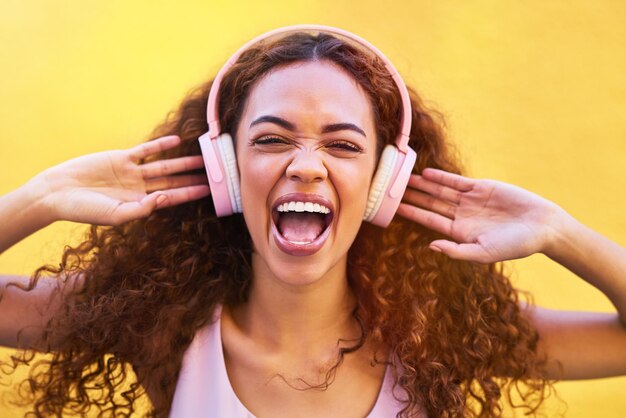 Musikalisches Gesicht und Geschrei mit einer schwarzen Frau, die draußen vor einem gelben Wandhintergrund Radio hört. Kopfhörer-Energie und Gesicht mit einer attraktiven jungen Frau, die zum Spaß Audio-Sound streamt