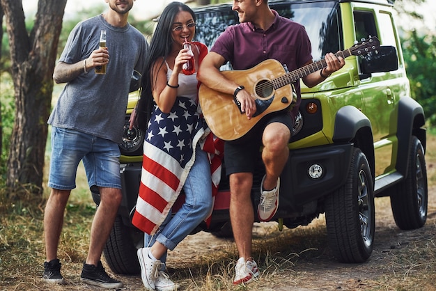 Musiker spielt ein Lied auf der Gitarre. Freunde haben ein schönes Wochenende im Freien in der Nähe ihres grünen Autos mit USA-Flagge.