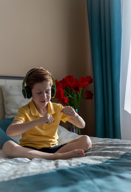 Musikliebhaber Freizeit und Unterhaltung Porträt eines rothaarigen Jungenkindes hört Musik über Kopfhörer, sitzt auf dem Bett und tanzt zufrieden vertikales Foto