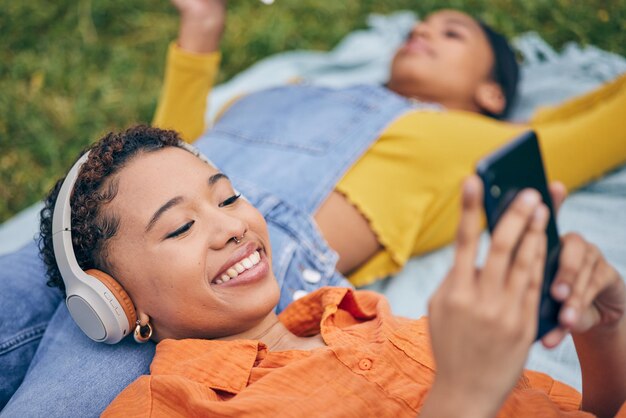 Foto musiktelefon und lesbisches paar entspannen sich beim picknick auf gras, technologie und streaming-service-app in der natur, handy, hören kopfhörer und glückliche lgbt-frauen auf einer decke im garten gemeinsam im park