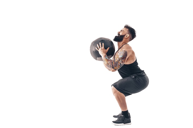 Muskulös tätowierter bärtiger Mann, der Fitnessgewichte ausübt Medizinball isoliert auf weißer Wand.