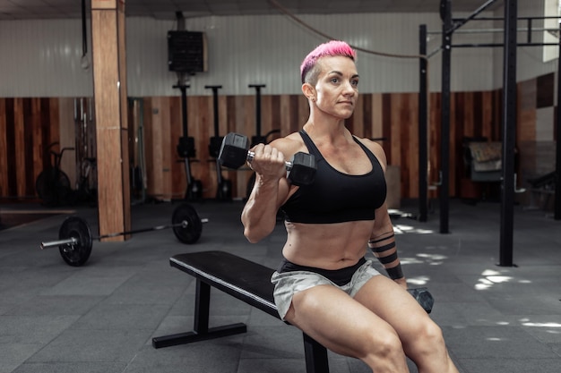 Muskulöse athletische Frau trainiert Bizeps mit Hanteln in ihren Händen, während sie auf einer Bank im Fitnessstudio sitzt.