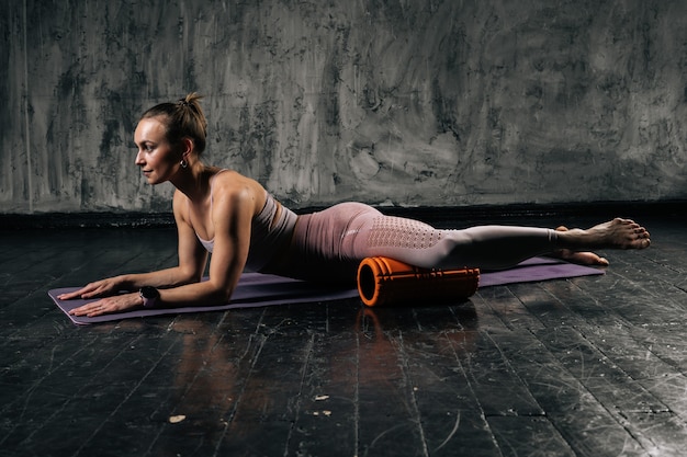 Muskulöse junge athletische Frau, die Fußmuskeln mit Schaumrollenmassagegerät massiert, das auf Yogamatte liegt.