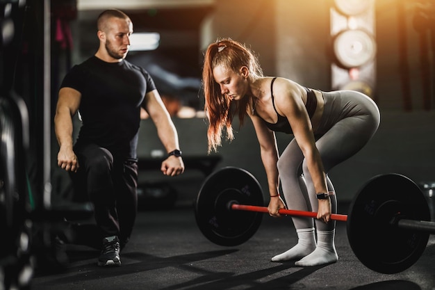 Muskulöse junge Frau, die mit Personal Trainer im Fitnessstudio trainiert. Sie macht Übungen mit Langhantel.