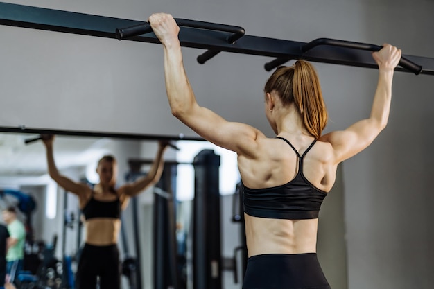 Muskulöser, athletischer Bodybuilder in Sportbekleidung, der sich im Fitnessstudio an einer horizontalen Stange vor dem Spiegel hochzieht. Rückenmuskeln eines sportlichen Mädchens, das sich strafft und sich im Spiegel ansieht