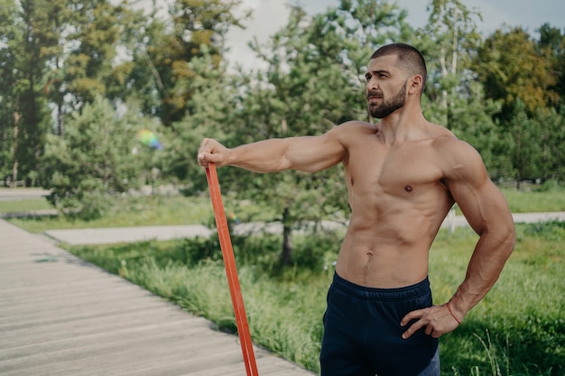 Muskulöser athletischer Mann hat Training mit elastischem Widerstandsband posiert mit hemdlosem muskulösem Körper, steht draußen, konzentriert in der Ferne. Sportler Sportler hat Training im Park. Gesunder Lebensstil