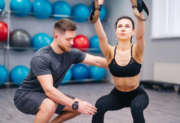 Muskulöser bärtiger Trainer, der attraktiven Frauen hilft, mit TRX im Fitnesscenter zu üben Sportliche Frau, die Übungen mit persönlichem Trainer im Fitnessstudio macht Nahaufnahme