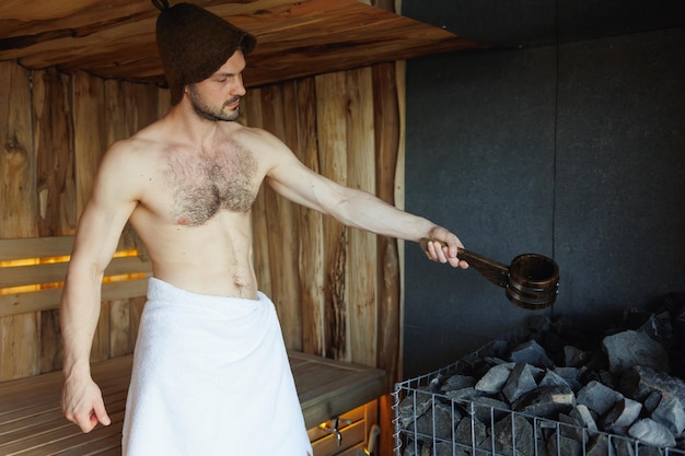 Muskulöser gutaussehender Mann gießt Wasser auf heiße Steine in der Sauna