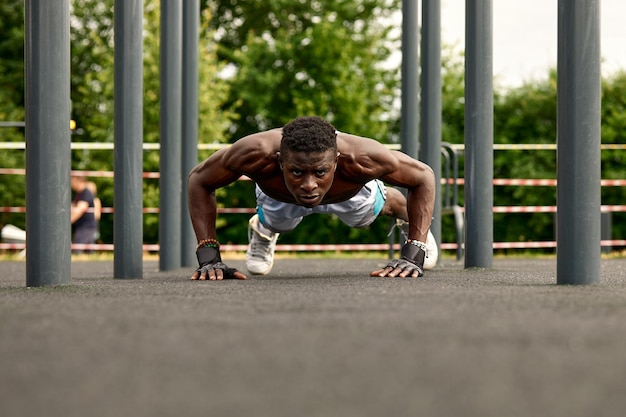 Muskulöser junger Mann, der auf dem Sportplatz trainiert Afrikanischer Mann, der zur Seite schaut, während er horizontale Stangenübungen macht Hemdloses männliches Modell, das im Freien trainiert