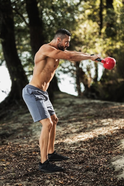 Muskulöser junger Mann mit nacktem Oberkörper, der Schwingübungen mit Kettlebell in der Natur macht.