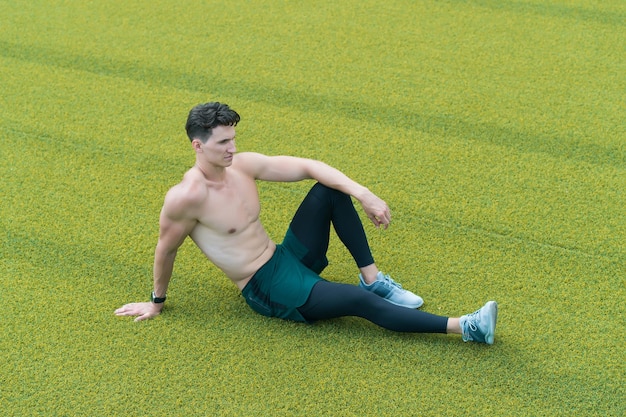 Muskulöser Kerl lässt auf Gras sitzen. Hemdloser Sportler, der sich nach dem Training ausruht. Kerl sitzt auf Gras. genuss ruhe entspannen. sportliches Lifestyle-Konzept. Entspannter sexy schulterfreier Mann, der auf Gras sitzt.