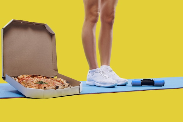 Muskulöser Körperbau passt zu Frauenbeinen, die auf einer Yogamatte in der Nähe einer Schachtel mit leckerer frischer Pizza und Hanteln stehen. Abnehmen und Fett bekommen Konzept. Fitness- und Diätkonzept.