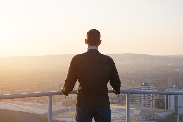 Muskulöser Mann in schwarzer Jacke hält die Hände an grauen Metallhandläufen und bewundert den Sonnenaufgang über dem Stadtbild und den Hügelsilhouetten von hinten