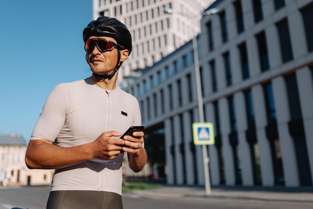 Muskulöser Mann in Sportkleidung, Helm und Brille, der zur Seite schaut, während er mit einem modernen Smartphone in den Händen auf der Straße steht Modernes Gebäude im Hintergrund