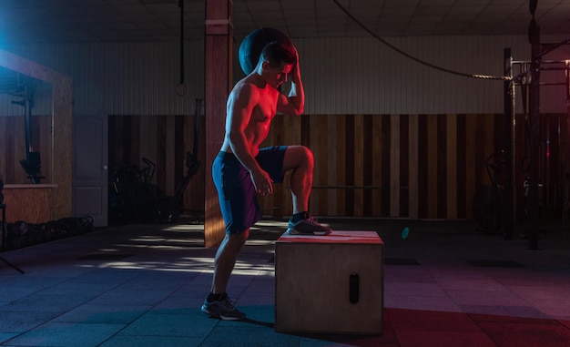 Muskulöser Mann trainiert Holzkiste mit einer schweren Tasche im dunklen Fitnessstudio Athletentraining in rot-blauem Neonlicht Gesunder Lebensstil Cross-Training