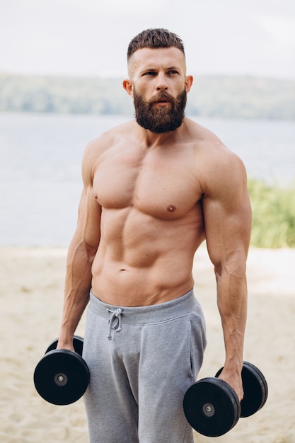 Muskulöser und bärtiger Mann mit Schnurrbart, der draußen Übungen mit Hanteln macht. Bodybuilding- und Outdoor-Sportkonzept