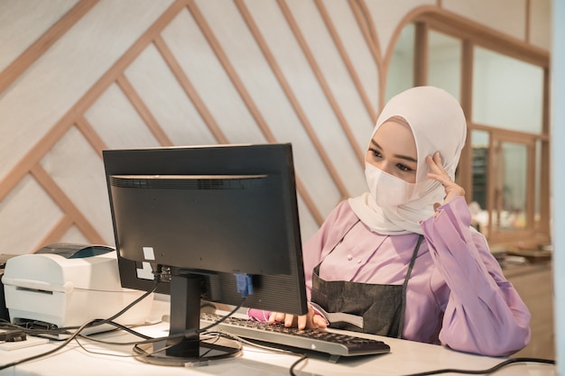 Muslimische asiatische Frau, die mit PC arbeitet, während sie medizinische Gesichtsmaske zum Schutz im Büro trägt