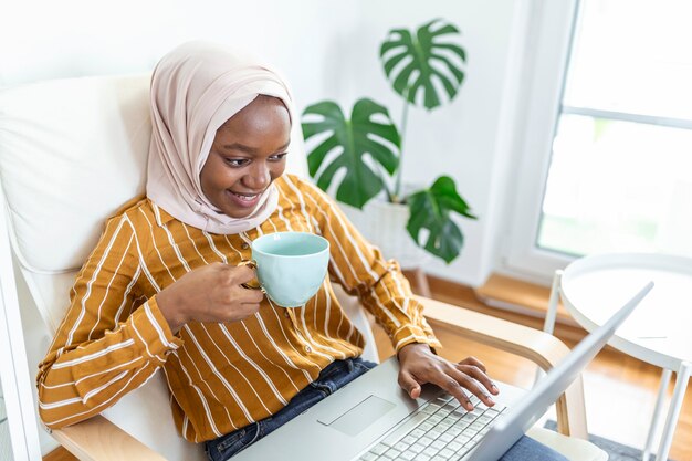 Muslimische Frau, die mit Computer arbeitet. Arabisch Junge Geschäftsfrau, die zu Hause an ihrem Schreibtisch sitzt, an einem Laptop-Computer arbeitet und Kaffee oder Tee trinkt. Muslimische Frau, die zu Hause arbeitet und Computer benutzt.