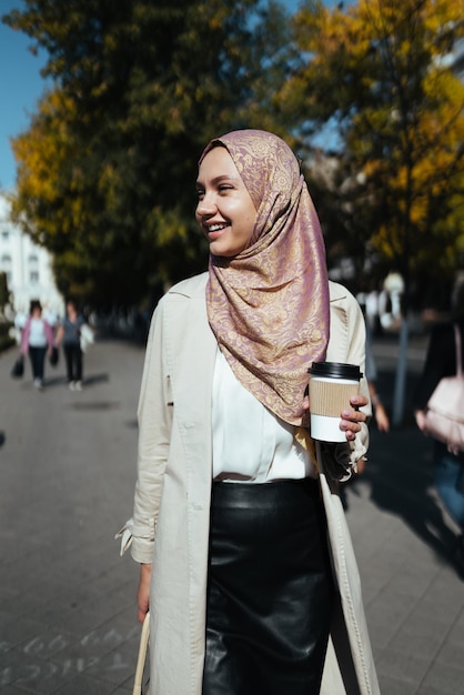 Muslimische Frau, die mit einem Glas Kaffee in der Hand die Straße entlang geht und lächelt