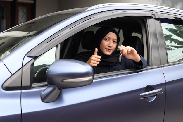 Muslimische Frau im Hijab fährt und zeigt Autoschlüssel aus dem Fenster, während sie den Daumen nach oben gibt.