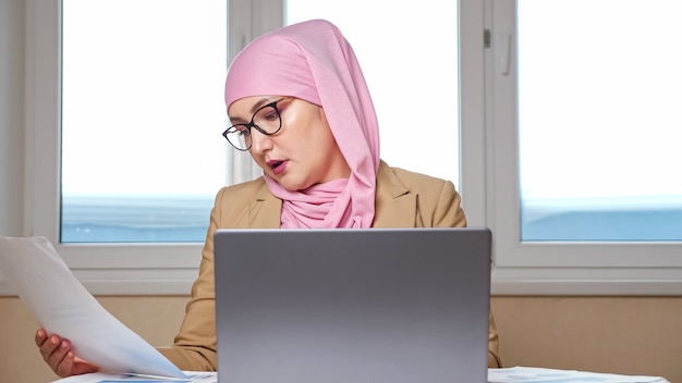 Muslimische Frau in rosa Kopftuch und Brille, die am Laptop und mit Dokumenten arbeitet.