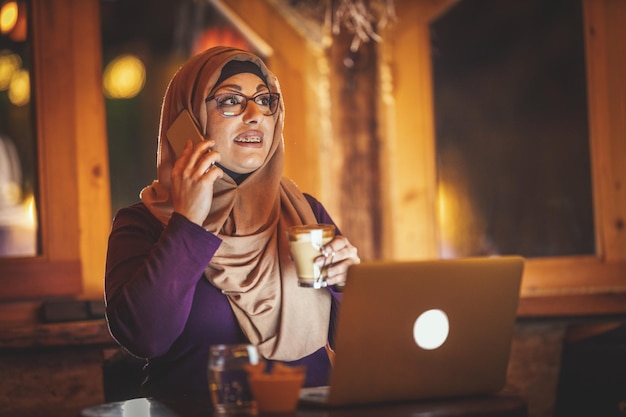 Muslimische Frau mittleren Alters, die Hijab trägt, sitzt im Café in städtischer Umgebung, trinkt Kaffee, spricht per Smartphone und arbeitet am Laptop.