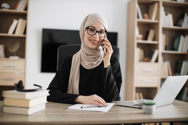 Muslimische Geschäftsfrau, die sich unterhalten, während sie am Tisch sitzt und an einem drahtlosen Laptop arbeitet