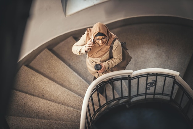 Muslimische Geschäftsfrau mittleren Alters mit Hijab steht auf der Treppe ihres Wohnhauses und nimmt ihr Smartphone in die Hand.