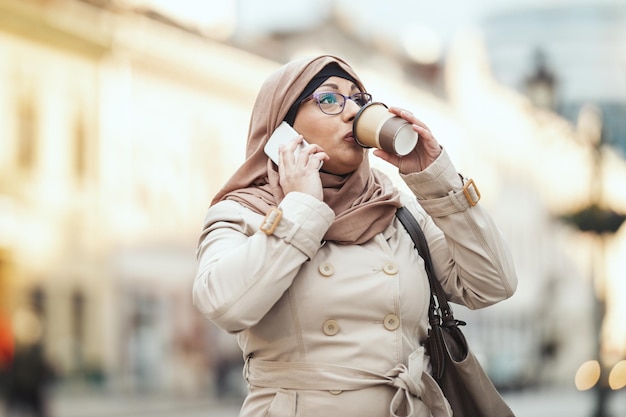 Muslimische Geschäftsfrau mittleren Alters mit Hijab und fröhlichem Gesicht steht in urbaner Umgebung, nimmt ihr Smartphone in die Hand und trinkt Kaffee.