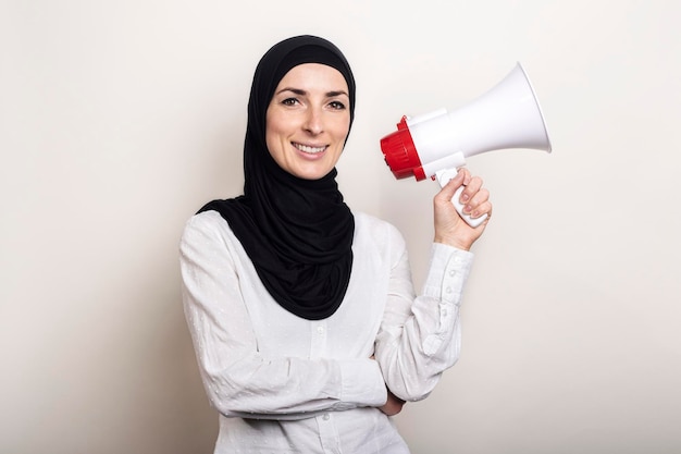 Muslimische junge Frau im Hijab hält ein Megaphon in ihren Händen und lächelt auf einem hellen Hintergrundbanner