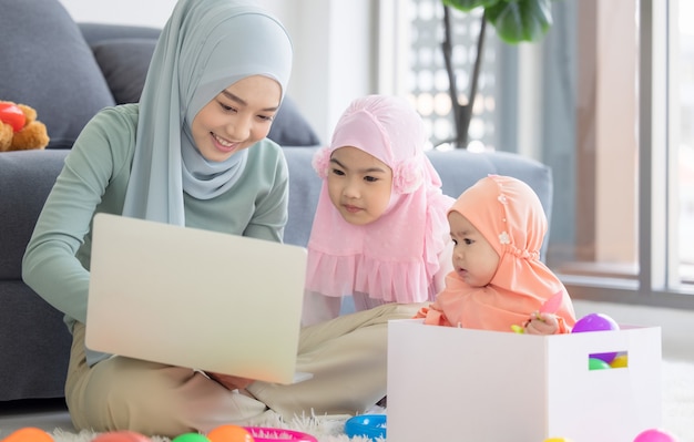 Muslimische Mutter, die mit Laptop und niedlichem kleinen Baby arbeitet, das Spielzeug im Wohnzimmer zu Hause spielt.