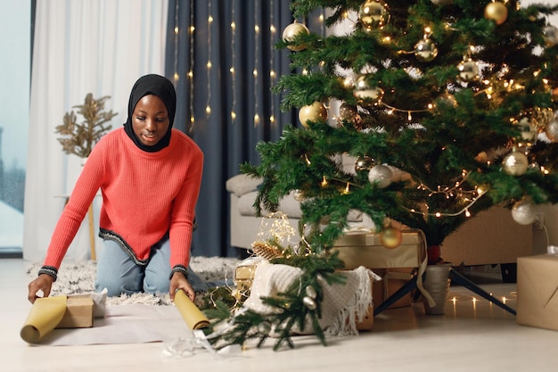 Muslimisches Mädchen im schwarzen Hijab, das in der Nähe des Weihnachtsbaums steht und eine Geschenkbox einwickelt