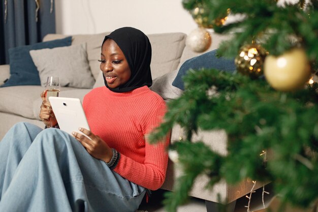 Muslimisches Mädchen in schwarzem Hijab sitzt mit einem Glas Champagner neben dem Weihnachtsbaum