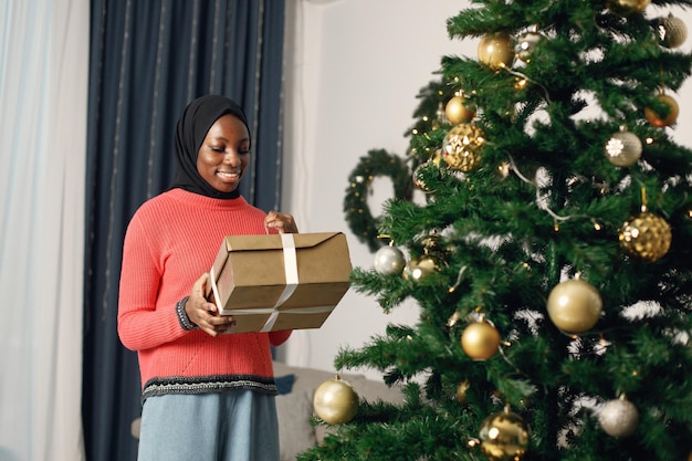 Muslimisches Mädchen in schwarzem Hijab steht neben Weihnachtsbaum mit Geschenkbox