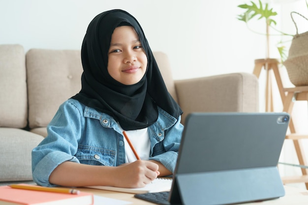 Foto muslimisches mädchen lernt online über das internet auf dem tablet, während im wohnzimmer zu hause morgens asiatische grundschulkinder sitzen und hausaufgaben schreiben konzept fernunterricht zu hause