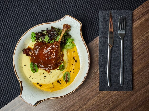 Muslo de pato con puré de calabaza, salsa de bayas y brotes de soja en una placa blanca.