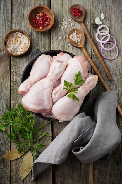 Muslos de pollo crudo en una olla de hierro fundido con especias y hierbas en madera vieja preparada para cocinar.