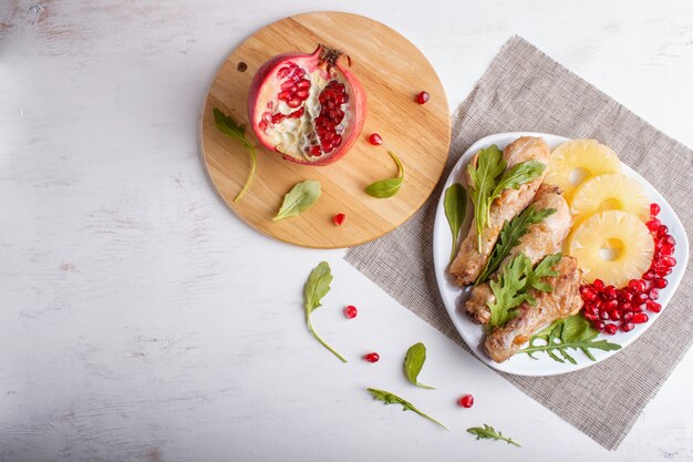 Muslos de pollo frito con semillas de rúcula, piña y granada en la superficie de madera blanca.