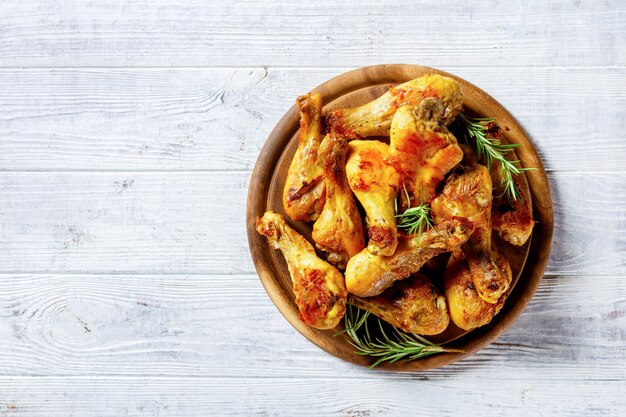 Muslos de pollo servidos en una tabla de cortar de madera sobre un fondo de madera blanca con ramitas de romero fresco, vista superior, primer plano