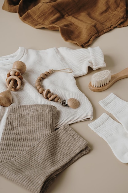 Musselin-Decke, Pullover, Leggings, Bürste, Socken, Schnullerhalter, neugeborene Babykleidung, Zubehör und Spielzeug auf neuralem beigen Hintergrund. Modische skandinavische Neugeborenen-Essentials