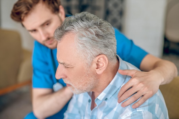 Mutloser grauhaariger kaukasischer Mann mittleren Alters, der von einem jungen, dunkelhaarigen, rücksichtsvollen Geriatriearzt unterstützt wird