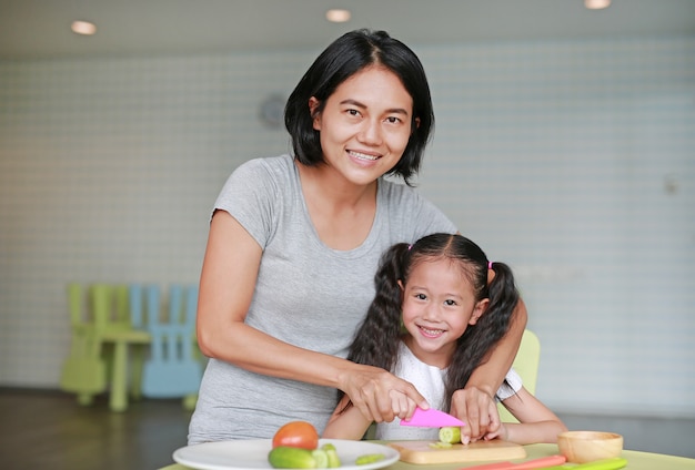 Mutter bringt ihren Kindern das Kochen bei. Schließen Sie herauf die asiatische Mutter und Tochter, die Gurkengemüse auf hackendem Brett am Spielzimmer schneiden.