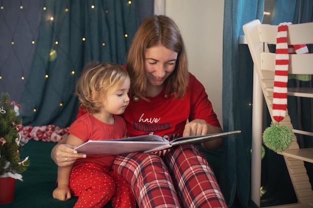 Mutter, die der kleinen Tochterfamilie ein Buch vorliest, sitzt auf einem weihnachtlich dekorierten Kinderzimmer