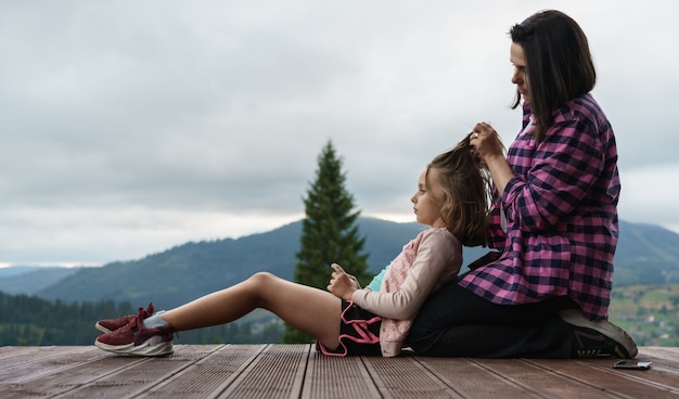 Mutter, die die Haare der Tochter im Freien flechtet, während sie auf der Terrasse mit Kopierraum sitzt