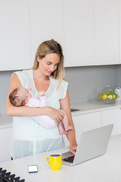 Mutter, die Laptop beim Tragen des Babys in der Küche verwendet