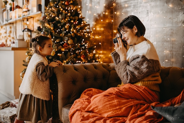 Mutter fotografiert ihre Tochter zu Hause neben einem Weihnachtsbaum